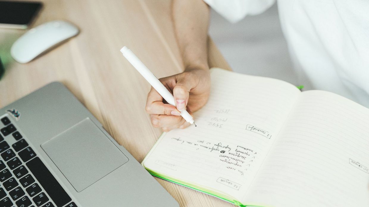 The SEO content writer job description: a hand writing in a notebook near a laptop, representing planning and freelancing